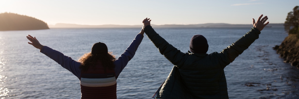 High above the shoreline, a couple holds hands with their arms raised and outstretched, celebrating the vast view of sun-dappled water and distant hills