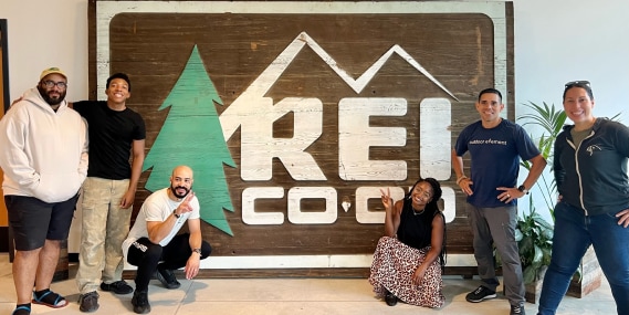 Six participants from the 2022 Navigate program co-hort gather around a wooden R E I Co-op sign inside the company’s headquarters.