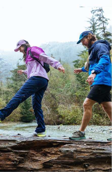 A group of hikers walk slowly, balancing on a fallen log along a river.