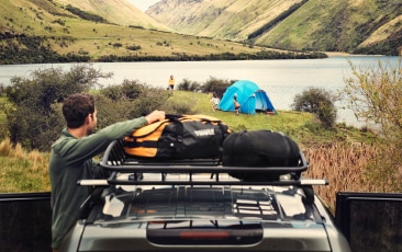 A person reaches into a Thule bag on their car rack, while looking out at their family’s campsite.