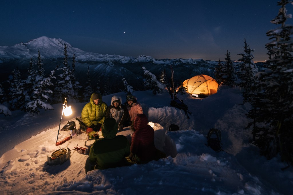 five campers huddled around a lantern on a cool night