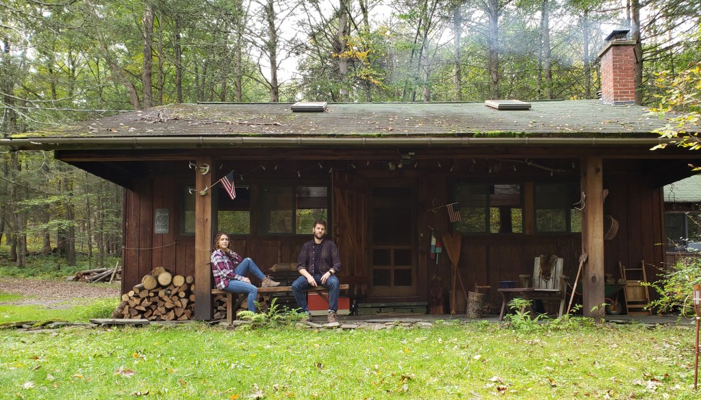 A couple outside of a brown wood cabin surrounded by greenery.