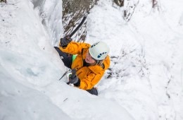 An ice climber in a helmet and bright yellow jacket swings her axe into a white frozen waterfall to move up the route.