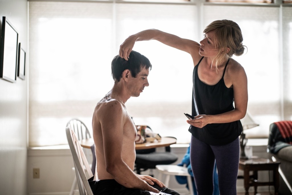 Alex Honnold sits in a chair while Sanni McCandless, standing, arranges his hair, holding scissors in one hand. 