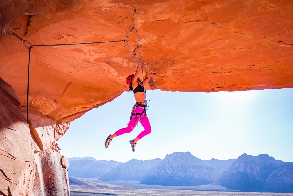 A climber dangles from a steep roof on a climbing route, with the desert landscape in the background.