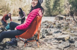 Camper with pink coat on sits in orange Flexlite chair while two people filter water in the background.