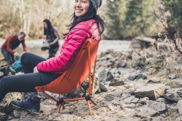 Camper with pink coat on sits in orange Flexlite chair while two people filter water in the background.