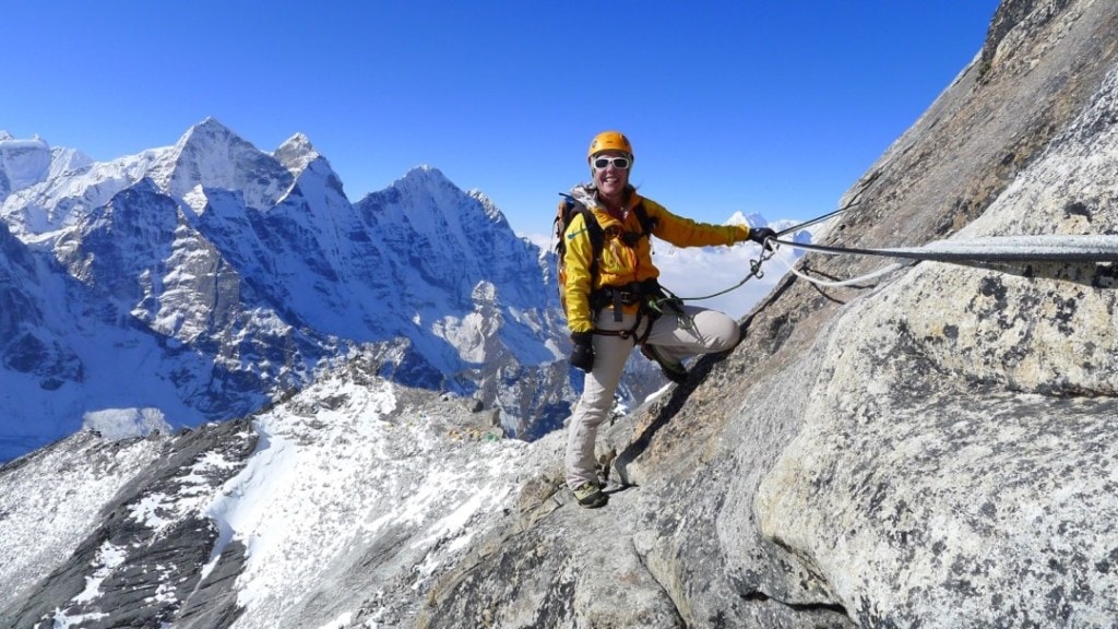 Dr. Monica Piris smiles in a yellow jacket and helmet while holding onto a rope on a rocky ridgeline. A collection of brightly colored base camp tents look very small in the background, with jagged, snowy peaks looming behind them.