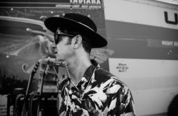 A black and white photo of G-Love facing away from the camera with a harmonica around his neck.