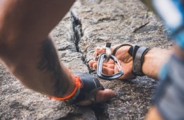 A rock climber wearing protective gloves holds onto a crack in a rock with one hand while inserting a cam for protection in the crack with the other hand.