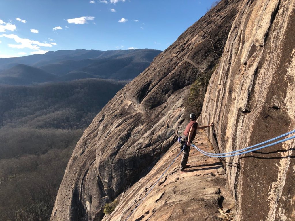 A barefoot rock climber stands on a curved ledge, looking over the edge to set up a rappel while tied into a blue rope.