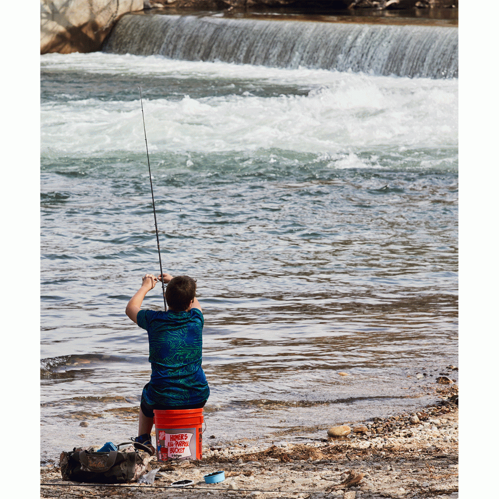 A young man uses a bucket as a seat while fishing along the Boise River. 