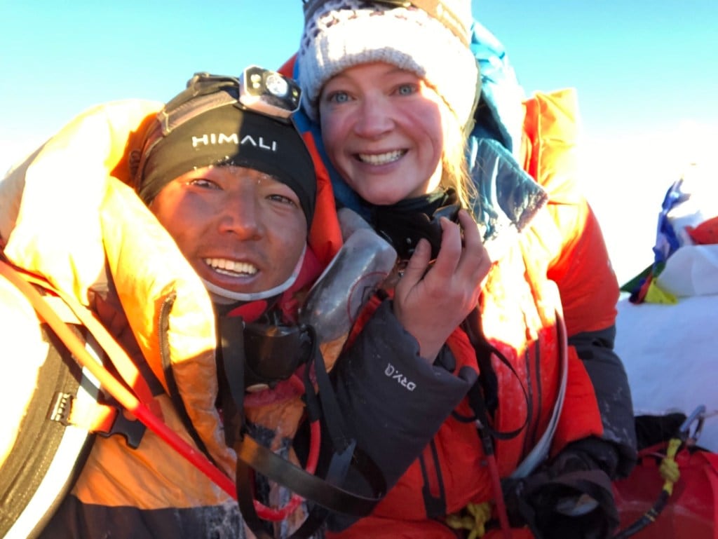 The author and her Sherpa guide remove their oxygen masks for a selfie on top of Everest with a bright blue sky in the background.