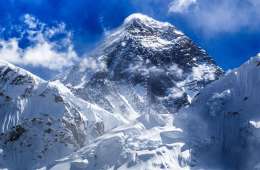 Mount Everest rises high against a blue sky with small, whispy white clouds. Chunks of ice scatter in the foreground.