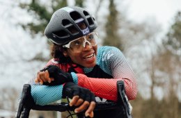 Ayesha McGowan leans on the handlebars of her bike while wearing a helmet, glasses, and a bike jersey