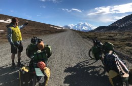 A person stands next to two bicycles packed full of gear on a dirt road and points to Denali in the distance