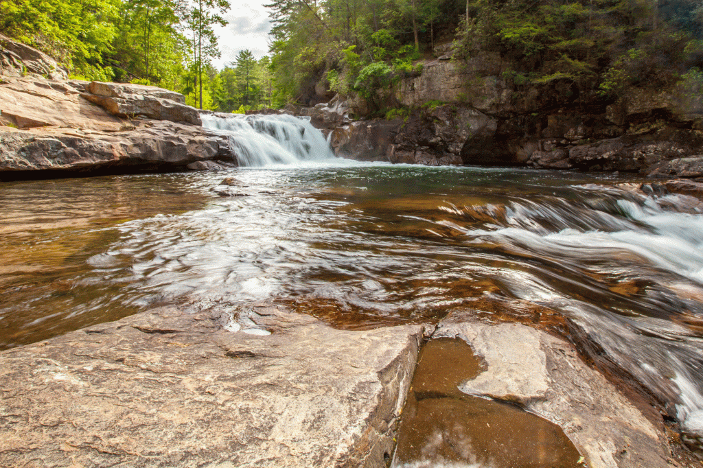 A rippling pool above light brown rocks beneath a rushing waterfall.