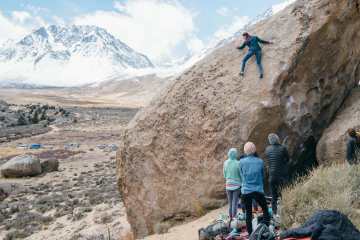 A rock climber makes a careful move on a large boulder while three people look on, spotting. A snowy mountain and a line of parked cars appear in the background.