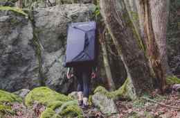 A lone climber walks a wooded path toward a grey boulder while carrying a bouldering pad.