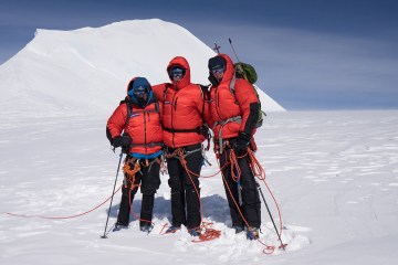 Three climbers in red puffy jackets stand, arms linked, tied into a rope on a snowy mountain.