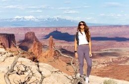 Melissa Urban standing at a viewpoint with the desert behind her. She's wearing a white tank top and grey athletic pants and is looking at the camera.