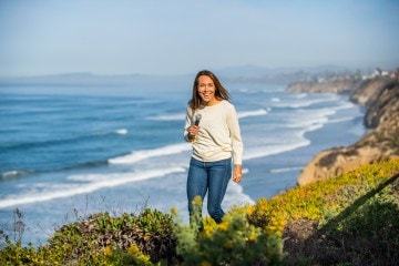 Shelby Stanger, host of the Wild Ideas Worth Living podcast, holding a microphone and looking at the camera with the ocean in the background.