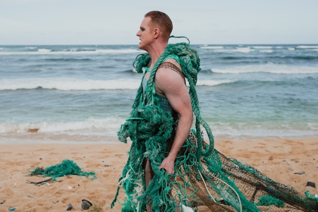 Pattie Gonia walks on a beach, adorned with a green net dress.