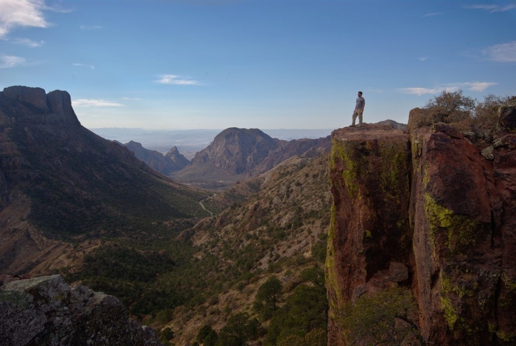 A person stands at the edge of a cliff overlooking a valley surrounded by mountains.