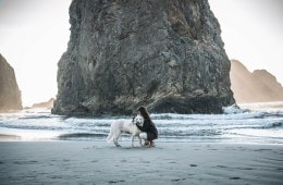 Ally is kneeling on the beach, with a rock stack behind her, while she hugs her white German shepherd around the neck.