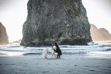 Ally is kneeling on the beach, with a rock stack behind her, while she hugs her white German shepherd around the neck.