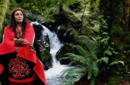 Harvest Moon, a Quinault native elder and storyteller, stands in front of a waterfall and ferns wearing a red cloak.