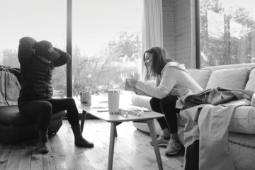 Two Arc'teryx designers are talking over a coffee table. One woman is pulling back her hair while the other woman holds a piece of fabric in her hand.