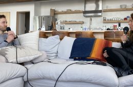 Mike Posner and Shelby Stanger are sitting on a white couch with microphones in their hands and headphones on, talking to each other during a podcast interview. This photo was taken pre-COVID.