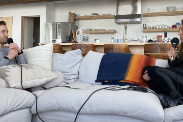 Mike Posner and Shelby Stanger are sitting on a white couch with microphones in their hands and headphones on, talking to each other during a podcast interview. This photo was taken pre-COVID.