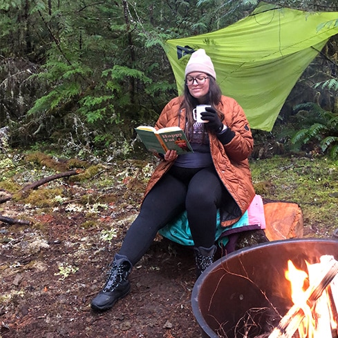 Woman reading a book by a campfire.
