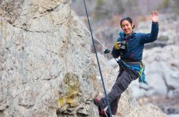 Smiling rock climber hanging from fix line holding camera and waving.