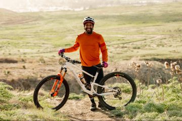 Eliot Jackson is posing behind his mountain bike with a green and golden field behind him. He's wearing an orange shirt and bike helmet and smiling at the camera.