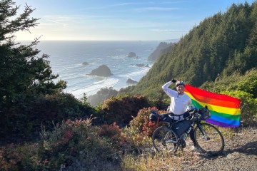 Mikah Meyer is standing on a trail in front of his bike holding a pride flag above his head with a background of trees and the ocean.