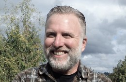 A close up shot of Cliff Barackman's face as he smiles at the camera. He's wearing a plaid shirt and has grey hair and a grey beard.