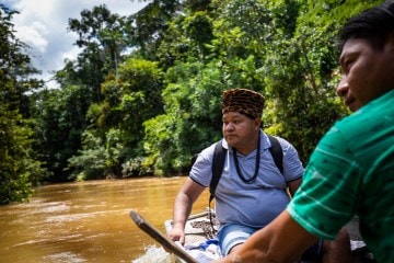 Two men from the Surui tribe sit on a boat in a river with the green rain forest behind them.