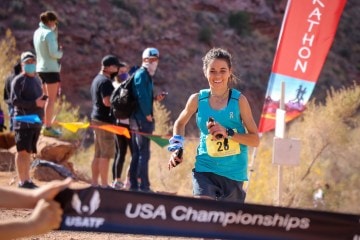 Allie McLoughlin, wearing a teal tank top is running on a dirt trail about to cross a finish line.