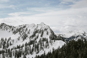 A snow capped jagged mountain ridge with pine trees, wispy clouds and blue sky on the horizon.