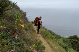 Diana Helmuth stands on a trail with the ocean in the background as she smiles back at the camera, a big backpack is on her back.