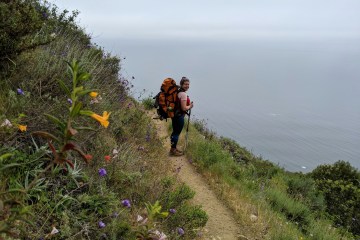 Diana Helmuth stands on a trail with the ocean in the background as she smiles back at the camera, a big backpack is on her back.