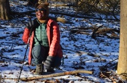 Lucienne Nicholson is crouched down in the snow with a hiking pole in one hand, laughing with a big smile on her face while looking at the camera.