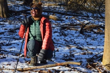 Lucienne Nicholson is crouched down in the snow with a hiking pole in one hand, laughing with a big smile on her face while looking at the camera.