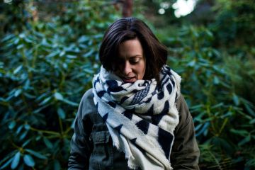 Steph Jagger is looking down at the ground with a white and black scarf around her neck a green shrug in the background.