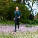 Woman with brown hair in a ponytail run across a field covered in pink flower petals. She's wearing a red and blue hydration vest and black athletic clothing.