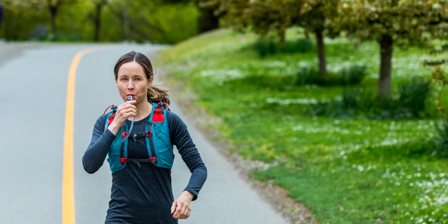 A woman with brown hair running down a road in a blue and red hydration vest, blowing water out of the tube of the hydration reservoir. 