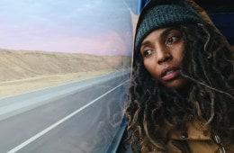 Brittany Washington is wearing a beanie and a hooded coat. She's leaning her head against a window with a road and desert outside.